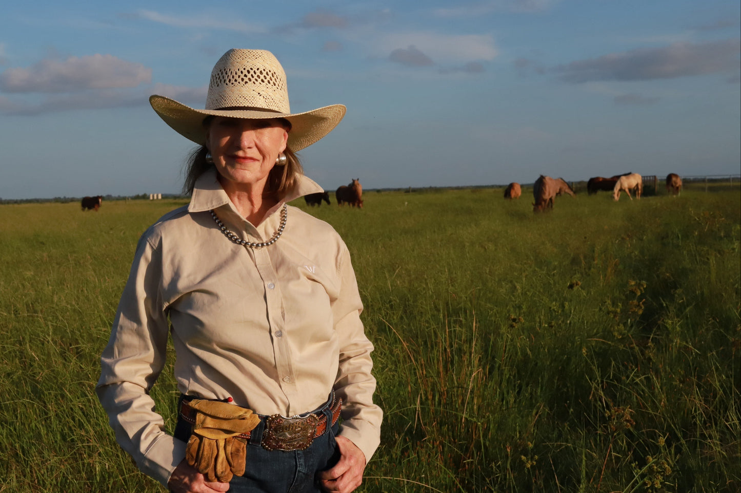 Woman in a cowboy hat and boots standing in a field with horses in the background