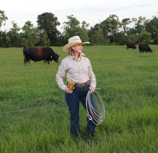 Woman in a cowboy hat and boots holding a lasso in a grassy field with cows in the background.